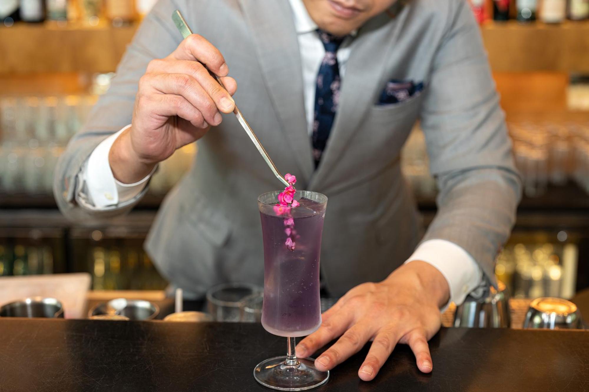 فندق Parkroyal Collection Pickering, سنغافورة المظهر الخارجي الصورة The photo shows a bartender in a stylish gray suit preparing a cocktail. He is holding a long, thin tool, possibly a stirrer or a tweezer, and is carefully placing a small pink flower garnish into a tall glass filled with a vibrant purple drink. The 